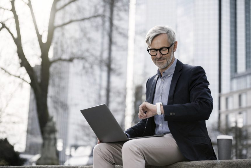 Grey-haired businessman working with laptop on wall in city looking at his smartwatch