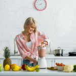 happy young woman preparing tasty nutritious smoothie in blender