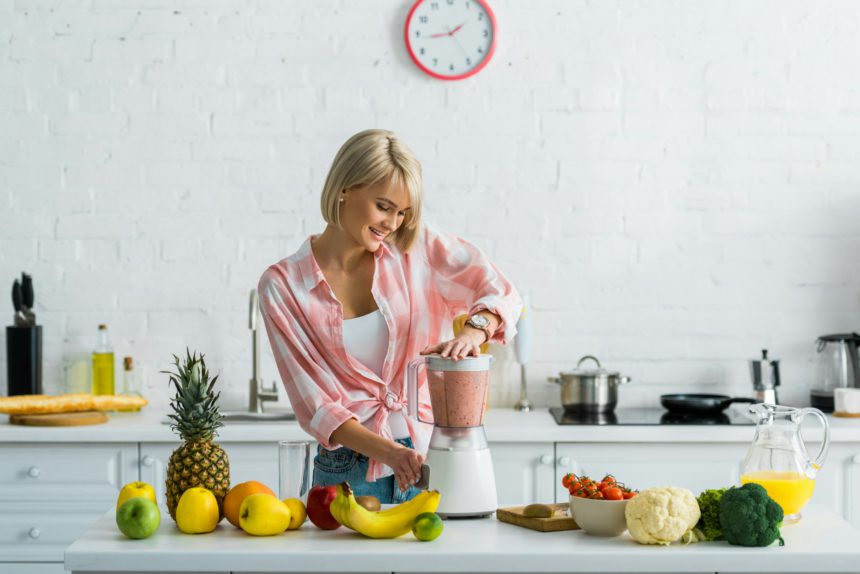 happy young woman preparing tasty nutritious smoothie in blender